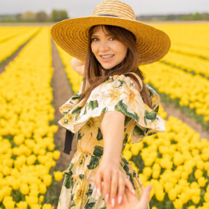 A woman smiling and holding hands wearing a hat and in a floral dress surrounded by yellow. tulip fields