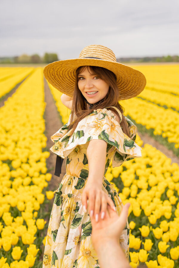 A woman smiling and holding hands wearing a hat and in a floral dress surrounded by yellow. tulip fields