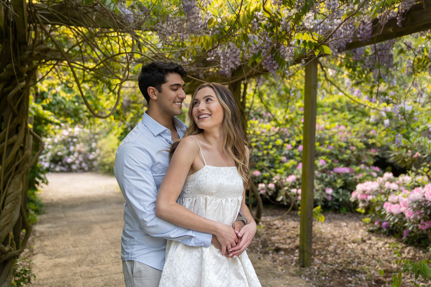 Engagement photo of a cute couple in a park surrounded by purple wisteria and pink flowers.