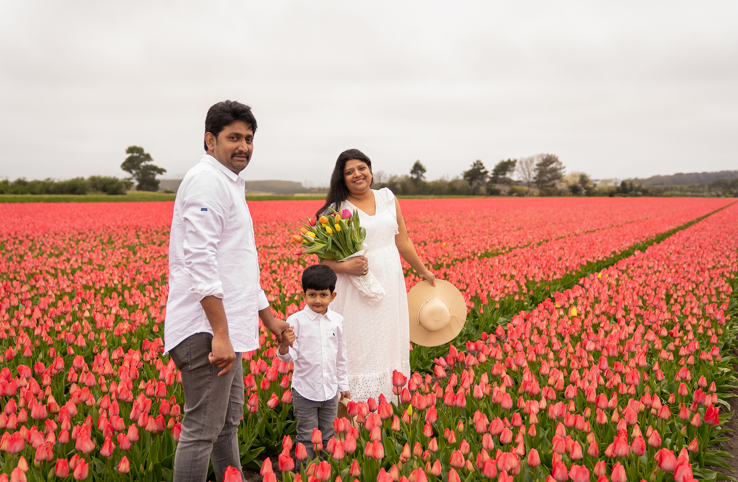 An Indian family of 3 smiling to the camera and wearing white clothing in the middle of red tulip fields.