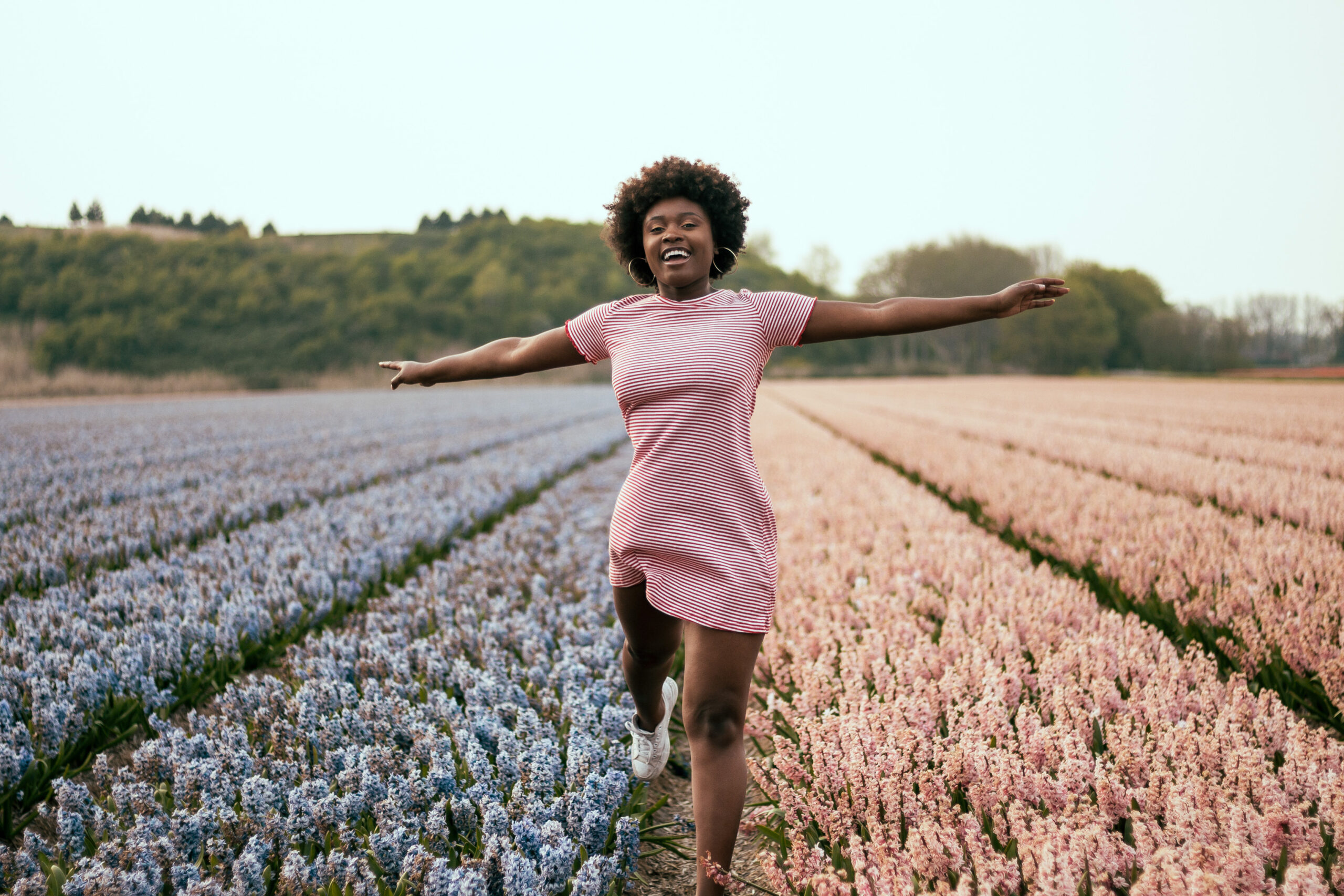 Afro-haired black woman smiles to the camera as she jumps through pink and purple flower fields in The Netherlands.