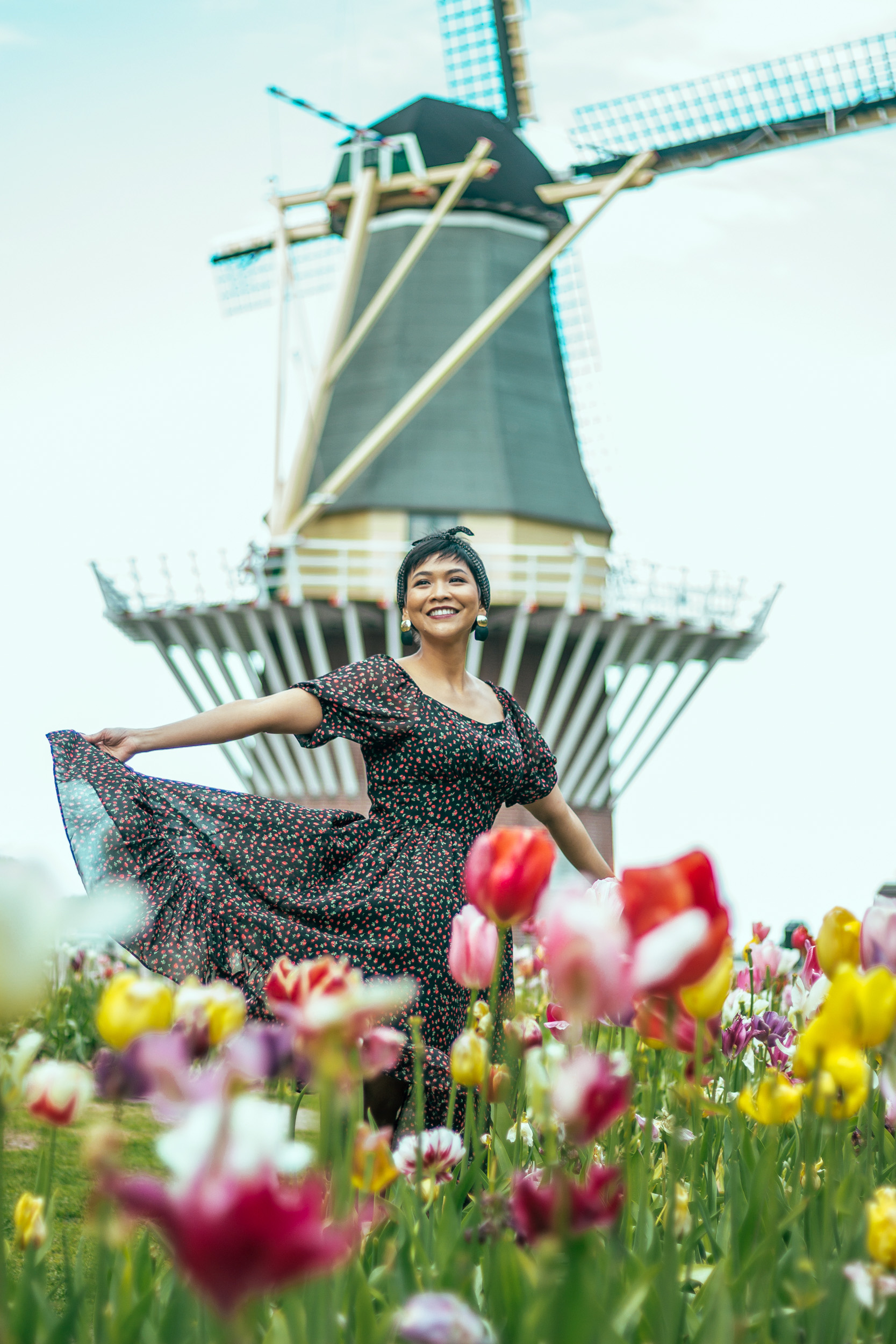 A woman is smiling and moving her dress surrounded by colorful tulips and a windmill in Keukenhof Gardens.