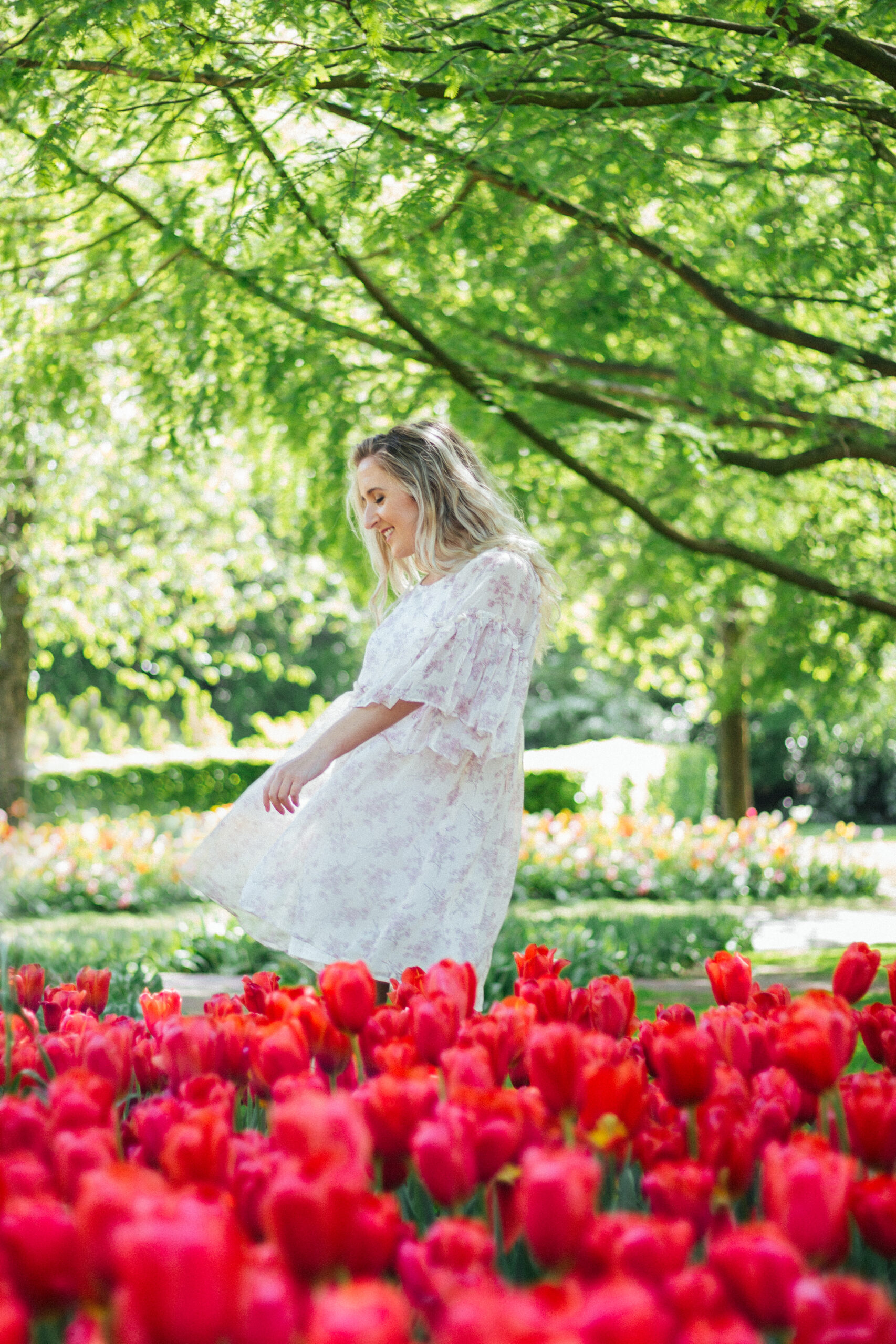 Blonde woman smiling at the Keukenhof tulip gardens surrounded by nature, trees, and red tulips.