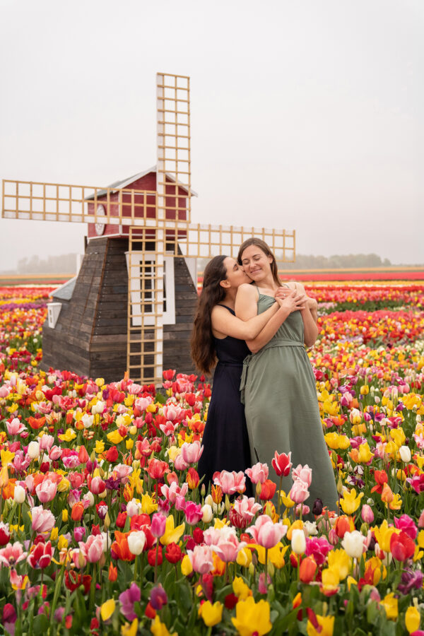 A lesbian couple who just got engaged is kissing in the cheek with colourful tulip fields and a windmill as backdrop.