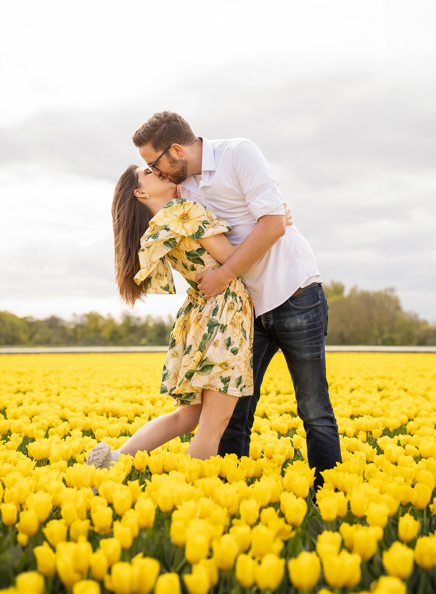 A couple kisses among yellow tulip fields while being photographed by a professional photographer.